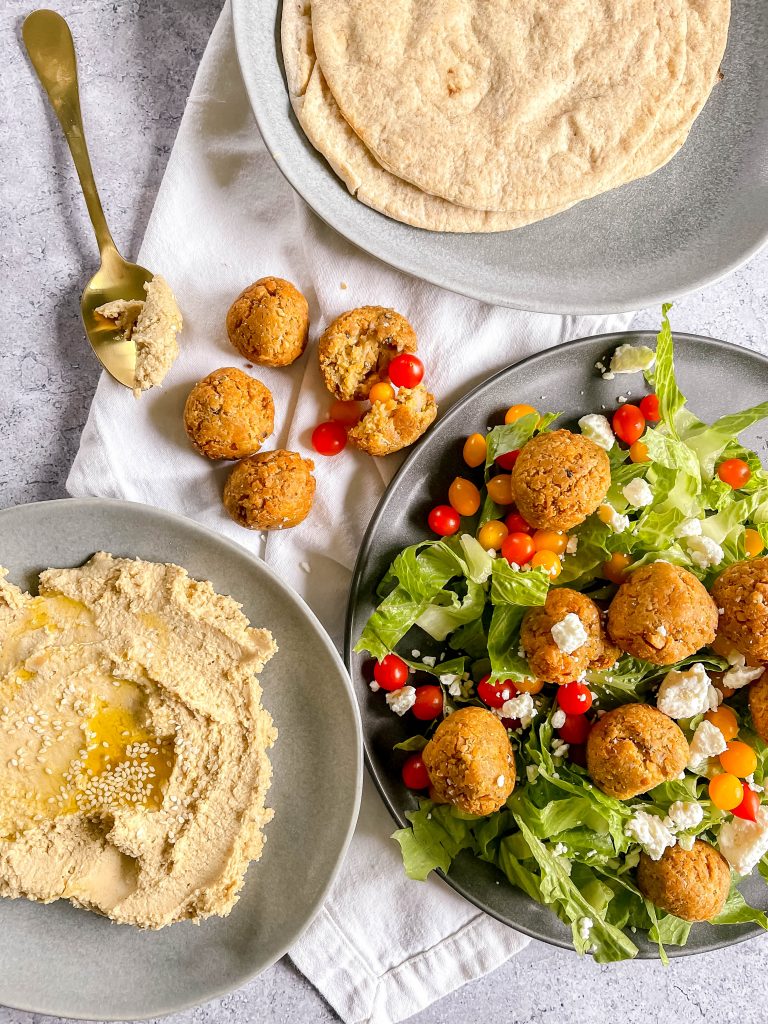 Three gray plates: one in the center right with falafel balls over salad, one to the left with hummus, one to the upper right with a stack of pitas. Gold spoon, cherry tomatoes scattered.