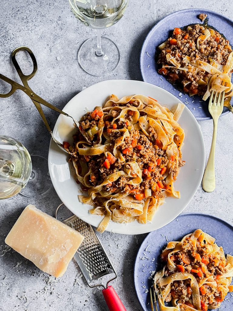 A big white plate full of bolognese with tagliatelle. Two other blue plates with helpings, a block of cheese, wine glasses, and forks. 