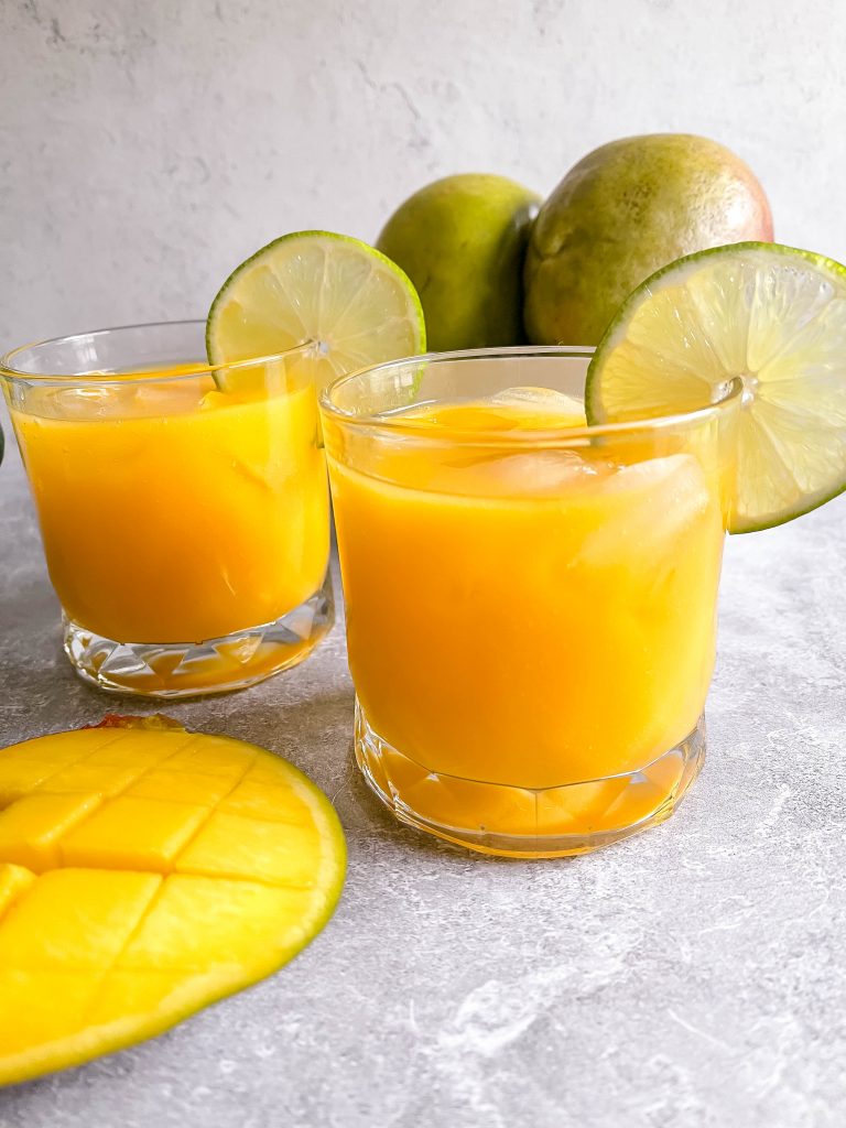 Two vibrant, orange margaritas in front of a carton of green mangos. 