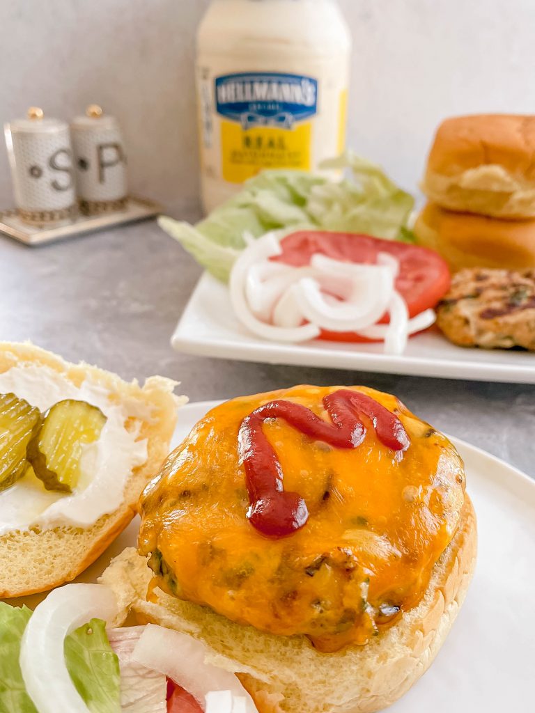 A juicy, cheesy burger up front with toppings, buns, and a jar of mayonaise in the back. 