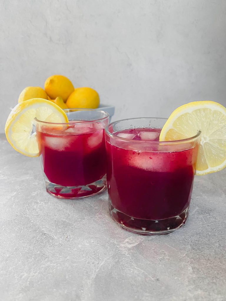 Two rocks glasses with a berry-colored drink, two lemon slices, and a carton of fresh lemons in the background.