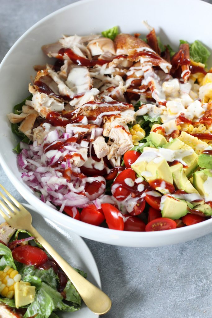A large white bowl with a bright salad consisting or lettuce, avocado, tomatoes, corn, red onion, chicken, bbq sauce and ranch!