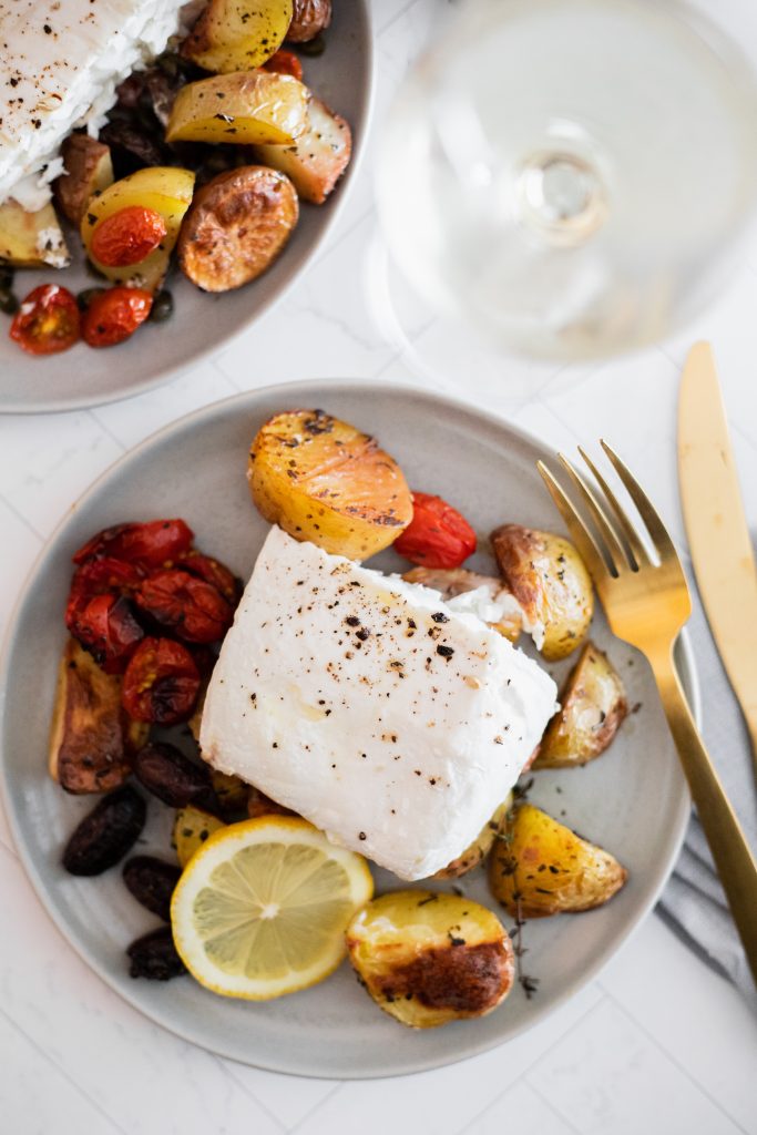 A gray plate with halibut, roasted potatoes, and vegetables. Gold flatware, and a glass of white wine.