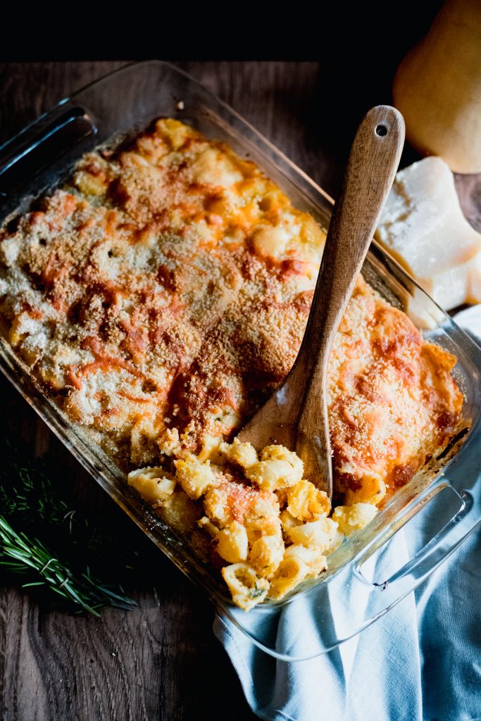 Baked macaroni and cheese in a clear baking dish with a wooden spoon, on a wooden table, a blue table cloth, with rosemary, cheese, and a butternut squash in the background.
