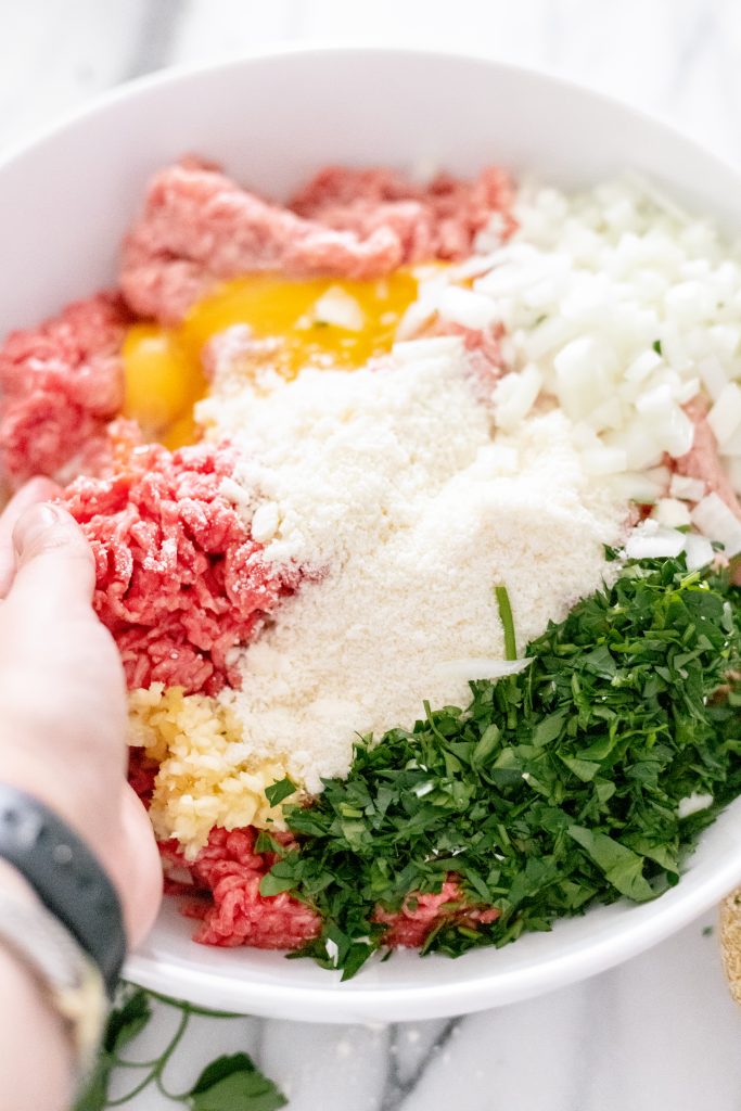 Ground beef, veal, pork, cheese, onion, parsley, eggs, and garlic in a bowl with a hand mixing the ingredients.