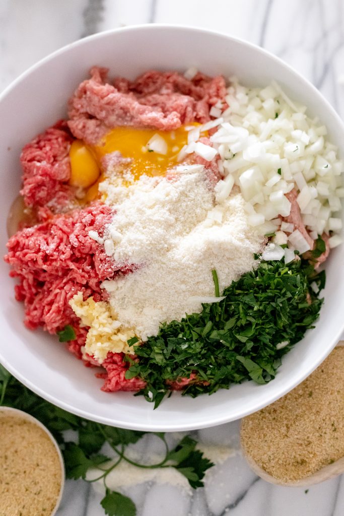Ground beef, veal, pork, cheese, onion, parsley, eggs, and garlic in a bowl.