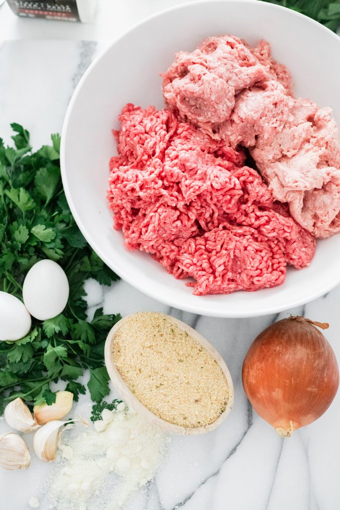 What ingredients do you need to make meatballs? A white bowl with ground veal, pork, and beef, parsley, onion, breadcrumbs, cheese, garlic, and herbs on a marble board. 
