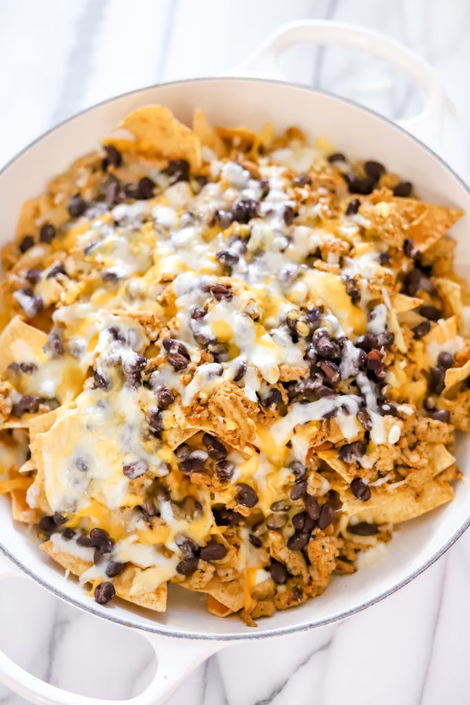 Cheesy nachos in a white pan on a marble backdrop - chips, beans, melted cheese, and ground turkey.