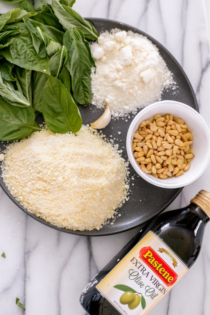 How to make basil pesto: olive oil, pine nuts, garlic, pecorino romano cheese, parmesan cheese, and basil on a gray plate with a marble backdrop.
