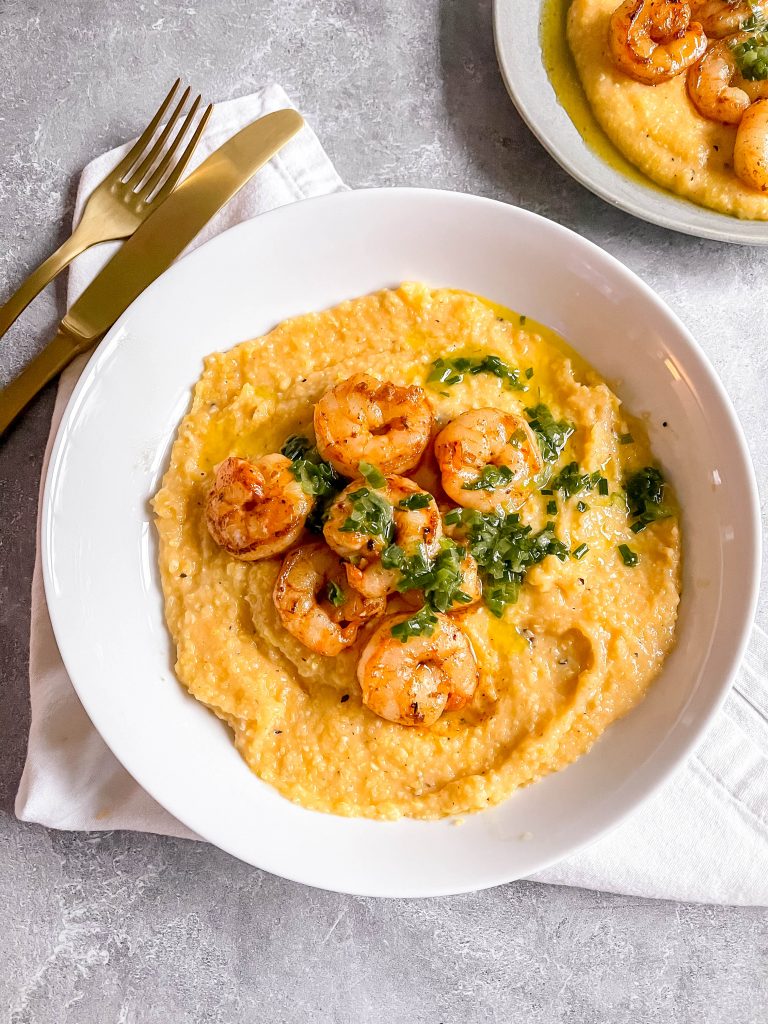 A bowl of cheesy grits topped with shrimp and green scallion oil. Stone background with gold flatware. 