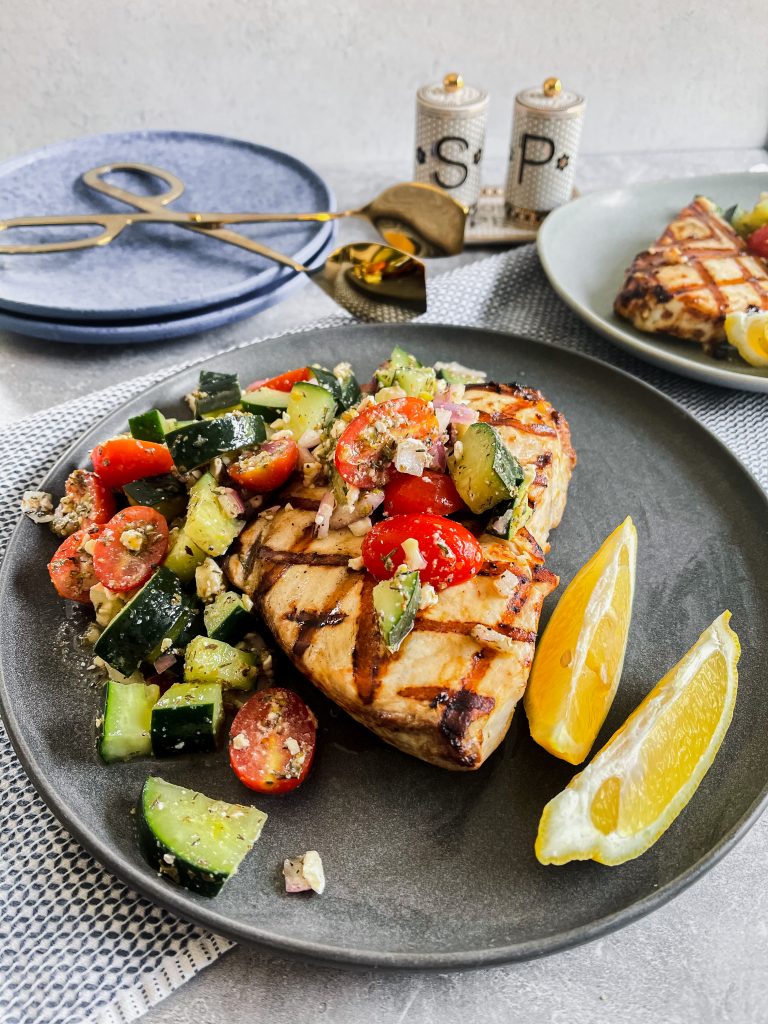 Grilled swordfish on a gray plate topped with greek chopped salad (tomatoes, cucumbers, feta, onion) with lemon. 