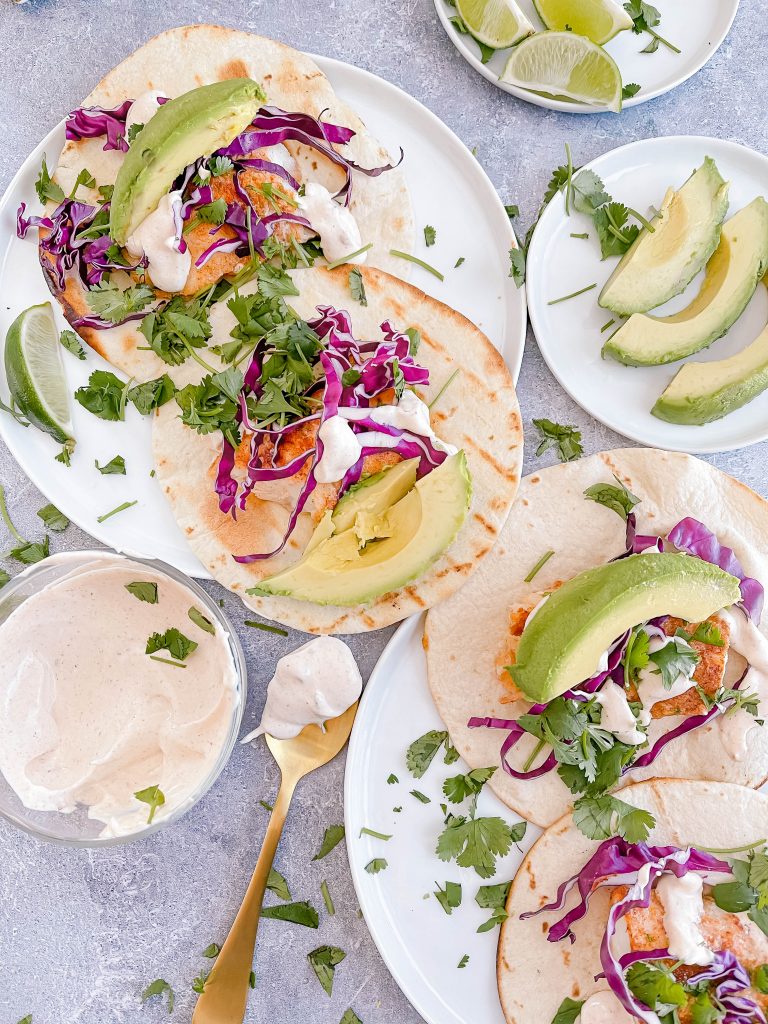 Two plates, each with two fish tacos: grilled tortilla, seasoned cod, red cabbage, cilantro, avocado, and crema. 