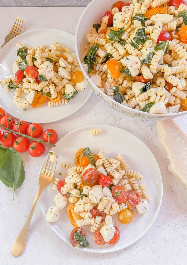 Two white plates and one big bowl full of pasta salad with tomatoes, cheese, and basil.