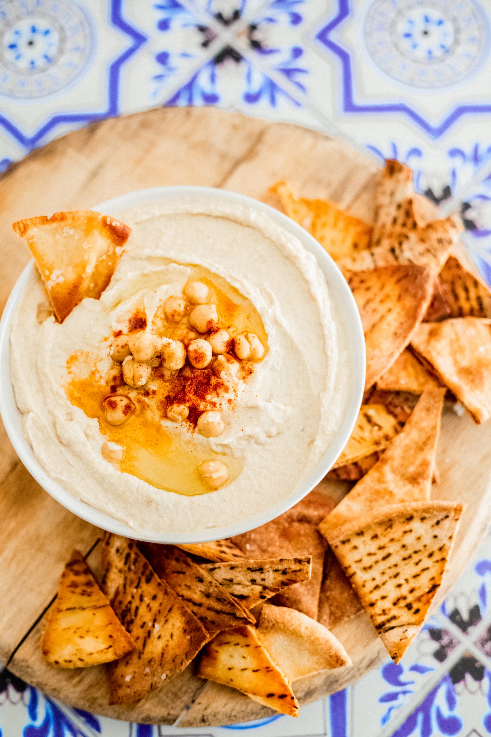 A white bowl of hummus with oli and chickpeas ontop. The bowl on a wooden circular board ontop of a blue tiled backdrop. There are pita chips surrounding the bowl.