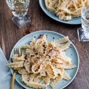 A blue plate on a wooden table with long pasta topped with pink peppercorns. Another plate of pasta in the background, two crystal wine glasses with white wine, and a golden fork.