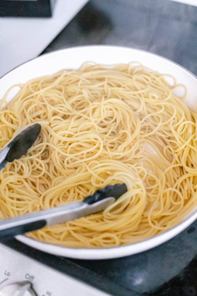 How to make spaghetti carbonara: spaghetti in a white pan with tongs on top of a stove.