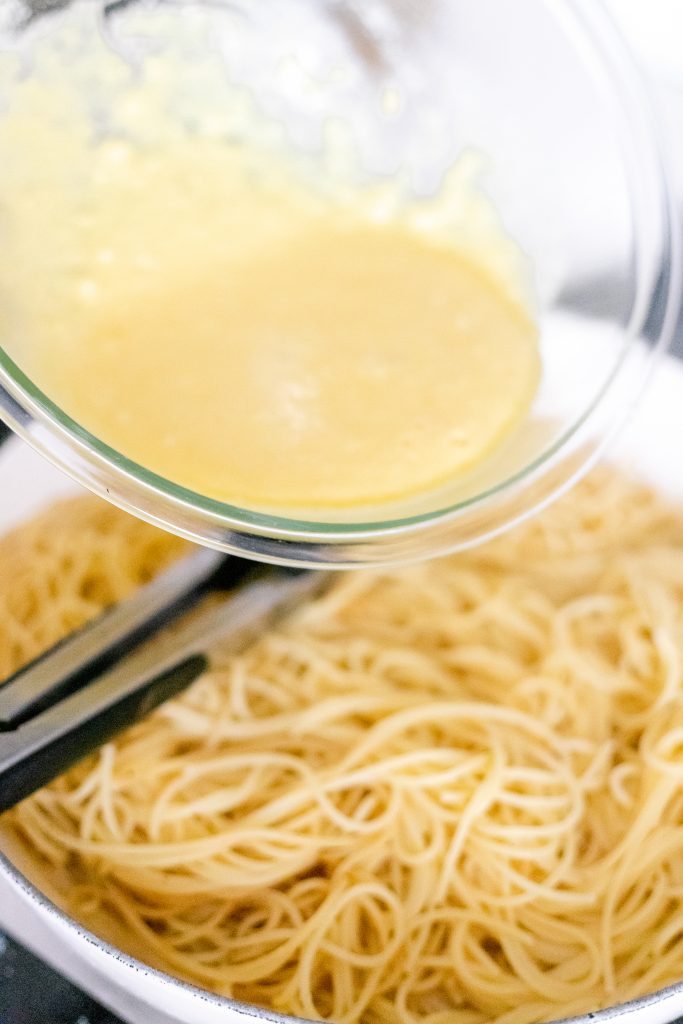 How to make spaghetti carbonara: a clear mixing bowl with a yellow sauce being poured into a pan full of spaghetti