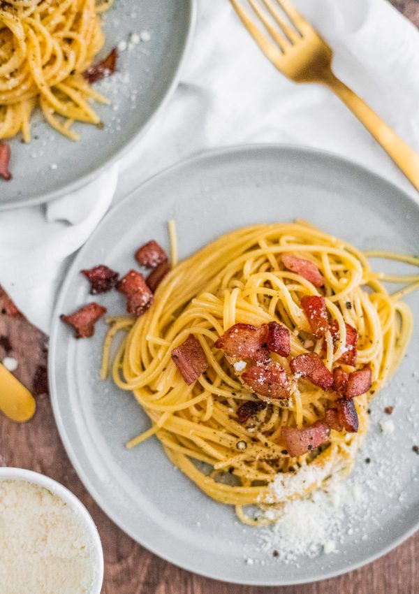 How to make spaghetti carbonara: two gray plates with spaghetti in a bright yellow sauce with crispy guanciale (it looks like bacon) and grated cheese and black pepper on top. A gold fork, a white napkin, and everything is on a wooden table.