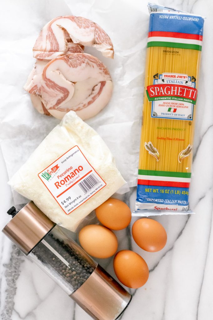 How to make spaghetti carbonara: ingredients on a marble backdrop: spaghetti, guanciale, pecorino romano cheese, 4 eggs, and a pepper mill.
