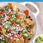 A big pan full of nachos with a side of guac on a wooden table