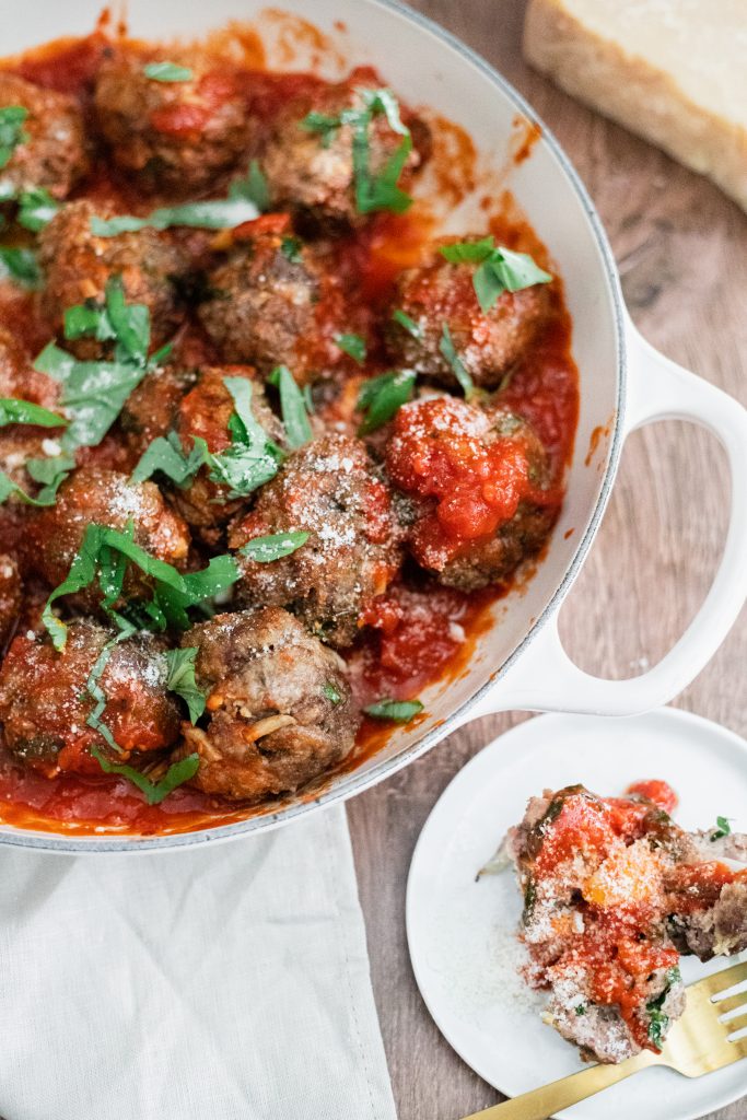 Italian meatball recipe: meetballs in a white pan with red sauce and chopped basil on a wooden table with a small white plate with a meatball and a gold fork