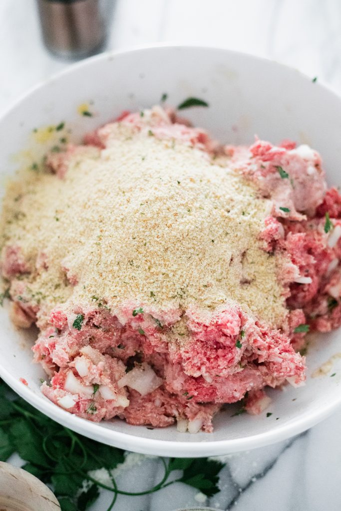 Mixed beef, veal, pork, cheese, onion, parsley, eggs, and garlic in a bowl with breadcrumbs ontop.