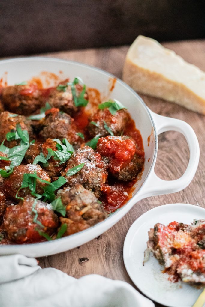 Meatballs in a white pan with red sauce and chopped basil on a wooden table with a small white plate with a meatball and a gold fork