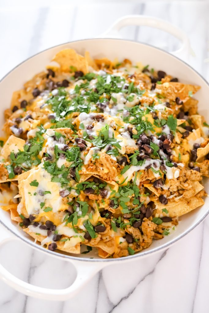 Cheesy nachos in a white pan on a marble backdrop - chips, beans, melted cheese, and ground turkey plus chopped cilantro