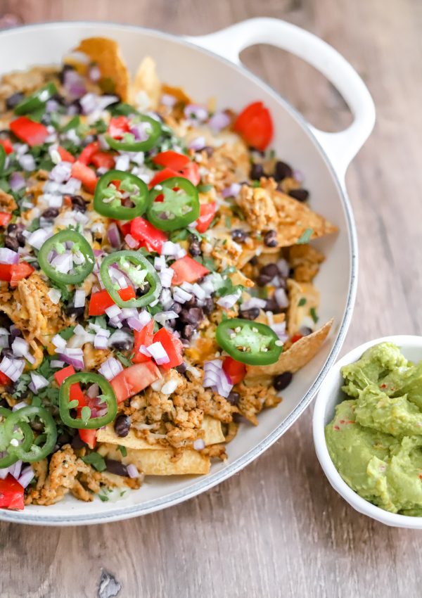 A big pan full of nachos with a side of guac on a wooden table