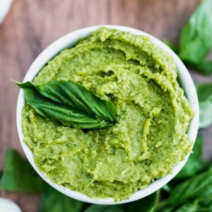 How to make basil pesto: vibrant green pesto in a white bowl on a wooden table surrounded by garlic and basil leaves.