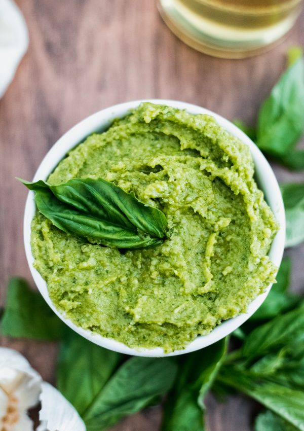 How to make basil pesto: vibrant green pesto in a white bowl on a wooden table surrounded by garlic and basil leaves.