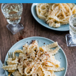 A blue plate on a wooden table with long pasta topped with pink peppercorns. Another plate of pasta in the background, two crystal wine glasses with white wine, and a golden fork.