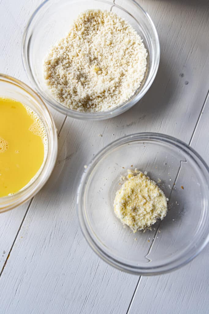 Overhead shot of three glass bowls on a white wooden table. In the glass bowls are a whisked egg (so all yellow), breadcrumbs, and a disc of goat cheese covered in breadcrumbs, respectively.