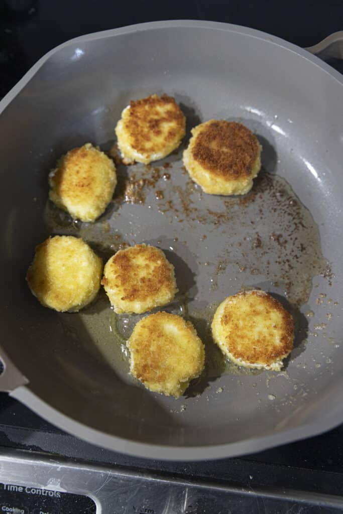 Overhead shot of goat cheese circles covered in breadcrumbs and fried, in a pan on the stove.