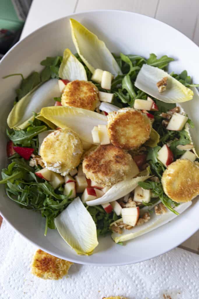 Overhead shot of chopped walnuts, diced red apple, endive leaves, and arugula, and fried goat cheese pieces, in a white bowl ontop of a white wooden table.