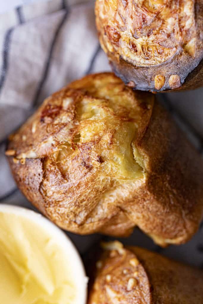 Overhead zoomed in shot of three popovers on the right hand side of the photo with a small bowl of butter in the bottom left corner and a linen gray and navy striped napkin in the top left corner.