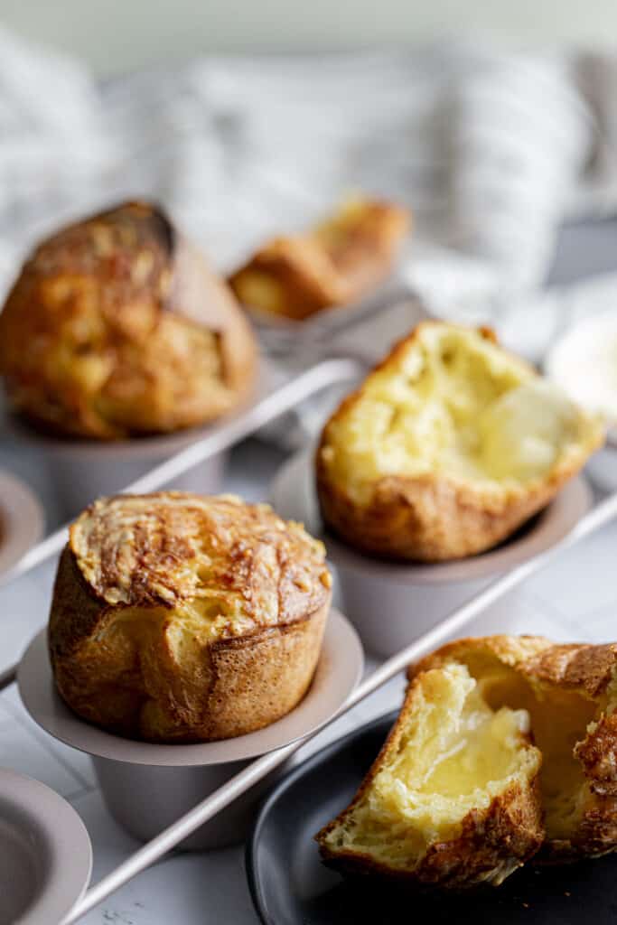 45 degree shot of popover pan filled with 3 popovers on a white marble countertop with a black plate with a ripped popover on it.