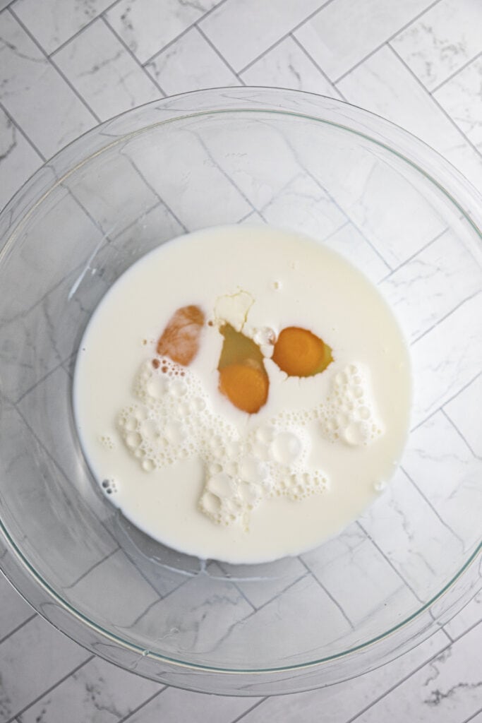 Overhead shot of milk and three cracked eggs with bright yolks in a glass bowl on a marble countertop.