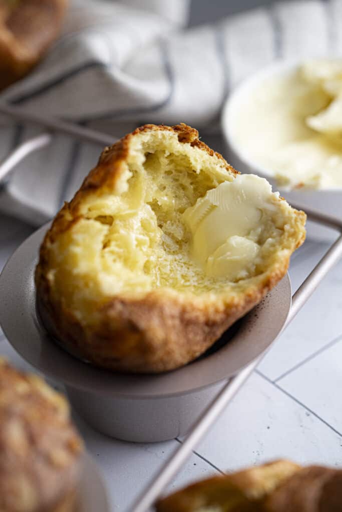 Zoomed in 45 degree photo of torn apart gruyere popover with melted butter inside in a popover pan on a white marble countertop.