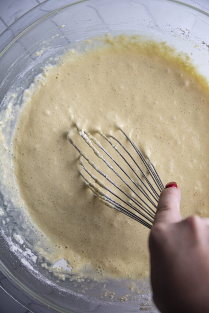 Overhead shot of hand whisking gruyere popover batter in a glass bowl.