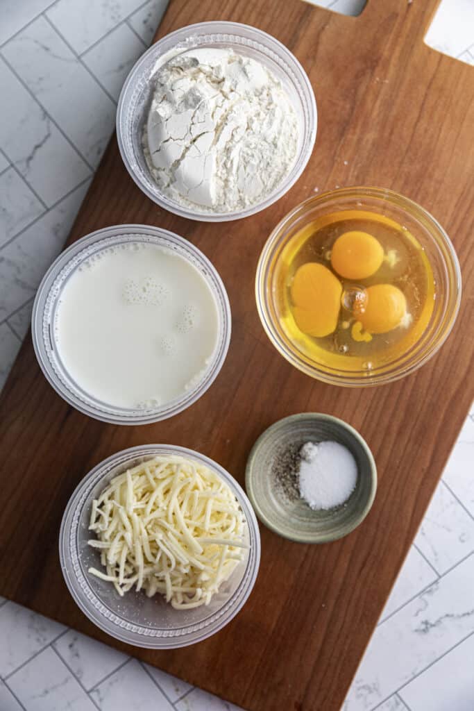 Overhead shot of ingredients for gruyere popovers: gruyere cheese, salt, pepper, milk, flour, eggs, all in individual bowls on a brown cutting board ontop of marble countertop .