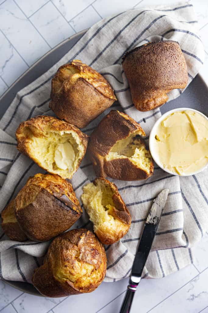 Overhead shot of 5 popovers (2 torn in half)on a gray linen towel with blue stripes with a butter knife and a small bowl of butter.