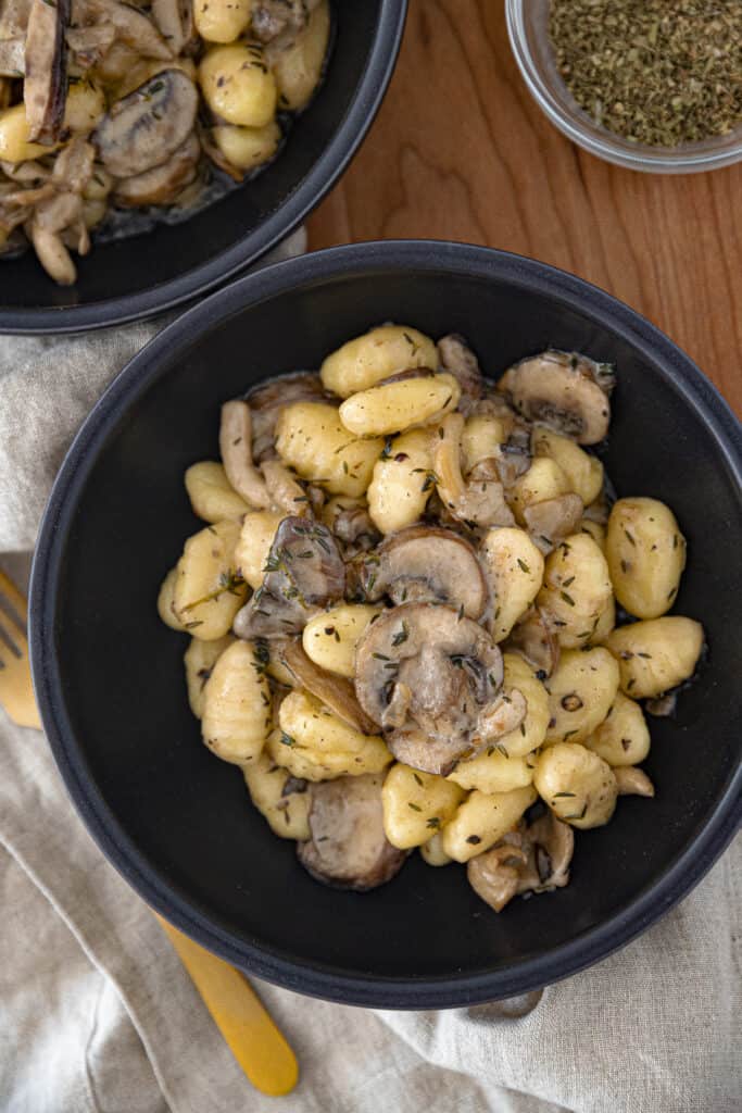 Two bowls full of gnocchi and mushrooms on a wooden cutting board with a gold fork