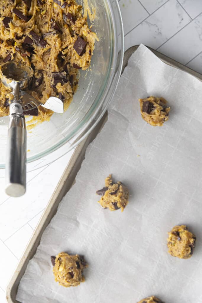 Overhead shot of rimmed baking sheet with parchment paper and raw cookie dough blobs, along with a bowl of cookie dough and scooper.