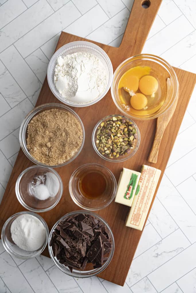 Overhead shot of ingredients for pistachio chocolate chip cookies: dark chocolate chunks, white sugar, brown sugar, salt, vanilla extract, butter, flour, pistachios, and eggs, all on a brown cutting board.