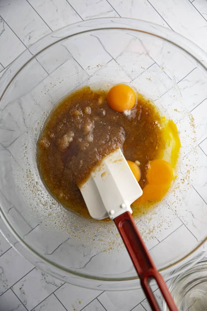 Overhead shot of brown butter and sugar mixed and three yolks unmixed in a glass bowl with a silicone spatula.
