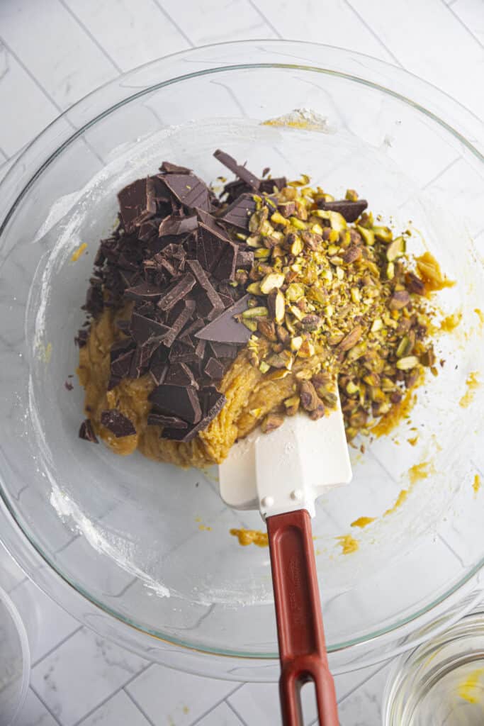 Overhead shot of cookie dough (unmixed) with a pile of chocolate chunks and a pile of chopped pistachios in a glass bowl with a silicone spatula.