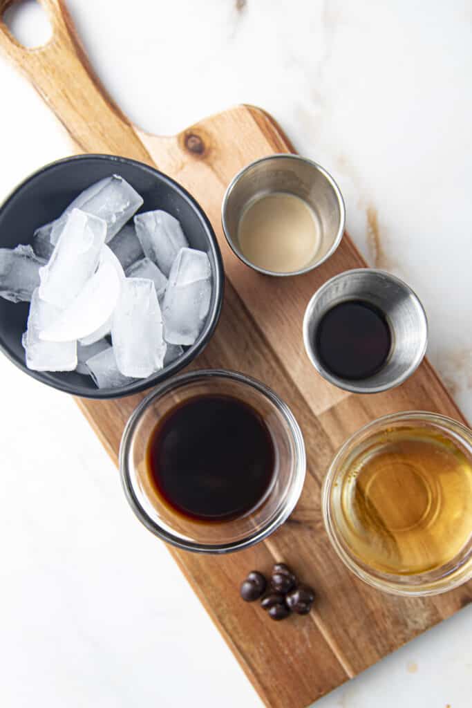 Ingredients for espresso martini with vanilla vodka: vanilla vodka, espresso, bailey's, kahlua, all in individual containers on a wooden board with chocolate covered espresso beans and ice, all on a marble countertop.