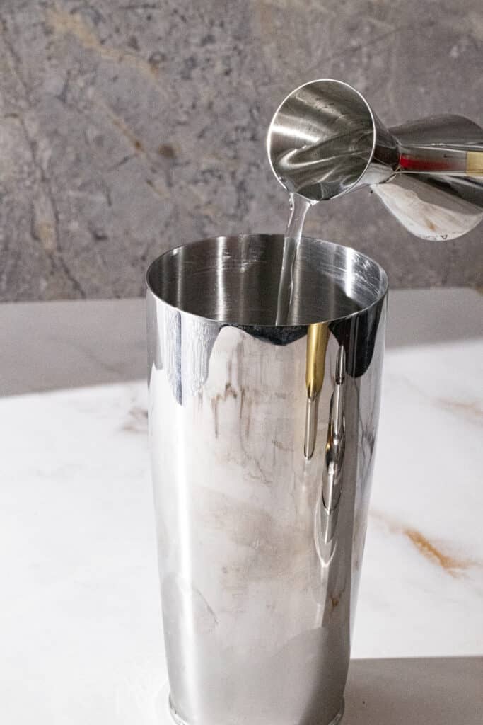 Silver cocktail shaker with vodka being poured into it on marble backdrop.