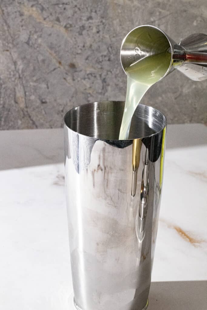 Silver cocktail shaker with limoncello being poured into it on marble backdrop.